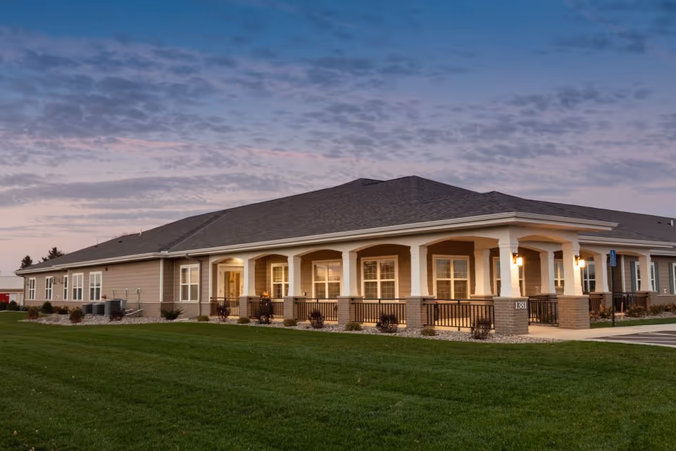 Exterior view of a single-story senior living facility building with a covered porch, multiple windows, and a well-maintained lawn under a partly cloudy evening sky.