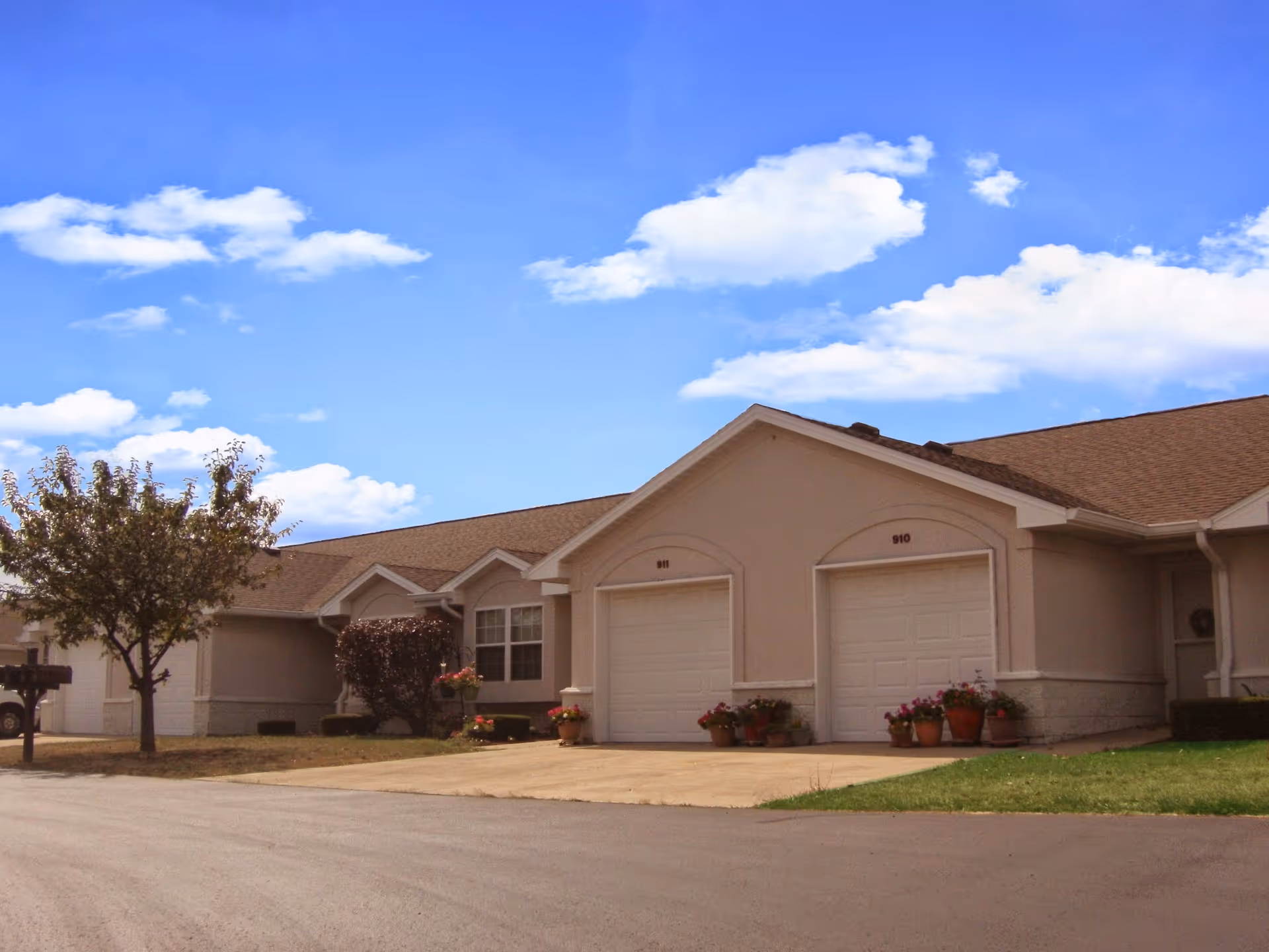 Exterior view of a single-story residential building with two garages numbered 910 and 911, a driveway, some potted plants near the garages, a tree, and a clear blue sky with scattered clouds.