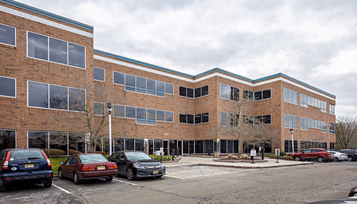 Three-story brick building with large windows and a parking lot under cloudy skies.