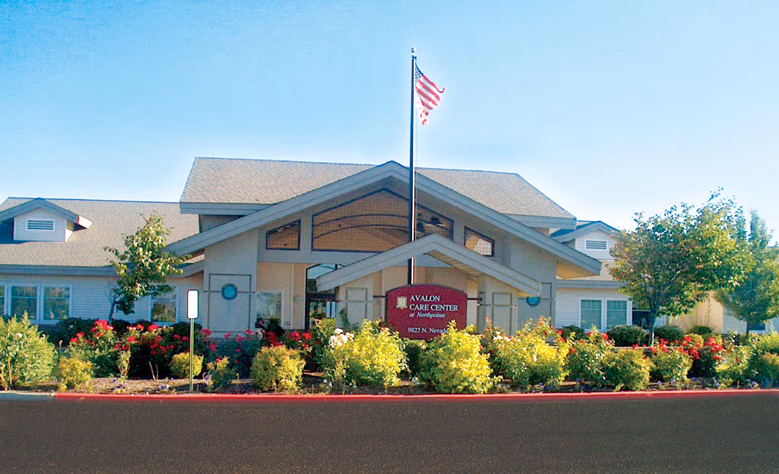 Front exterior view of Avalon Care Center at Northpointe building with a peaked roof, an American flag on a flagpole, and a landscaped garden with bushes and flowers in front.