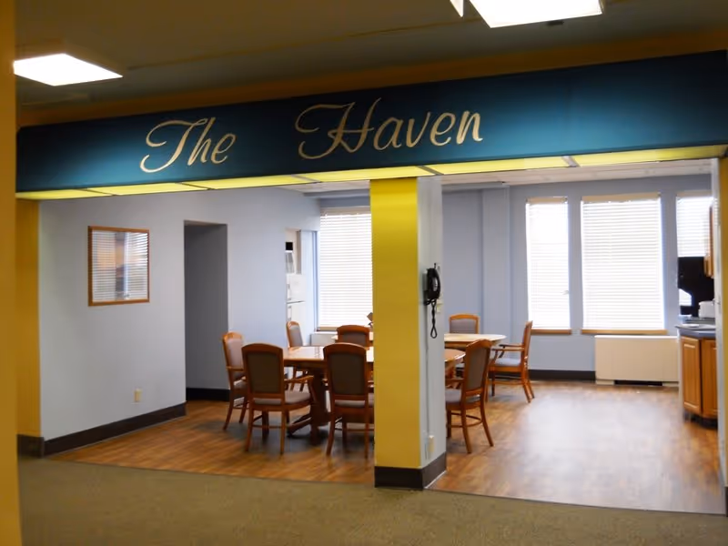 Interior dining room with round tables and chairs beneath a blue sign reading The Haven.