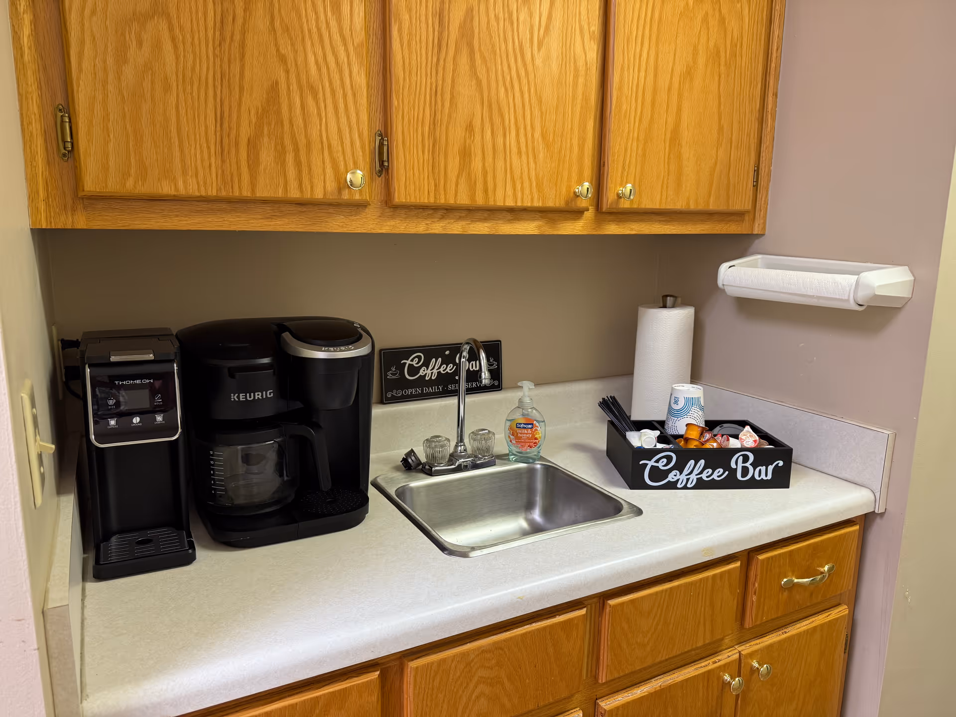 Coffee station with Keurig, coffee maker, sink, and paper towels in a kitchenette.