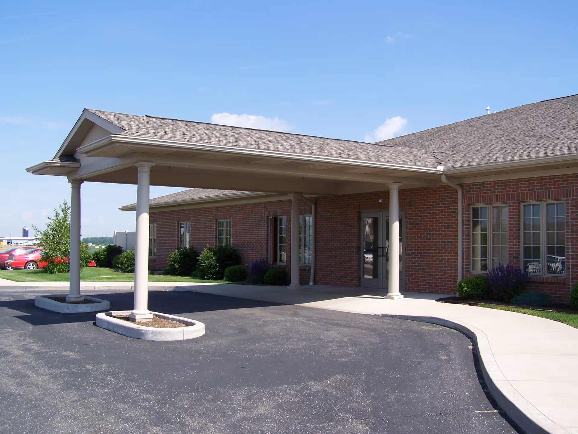 Brick facility entrance with a covered porte-cochère and parking lot under a blue sky.
