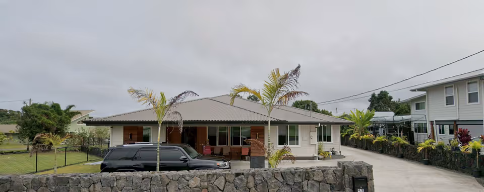 Single story residential senior facility with stone wall, palm trees, a driveway, and a black van.