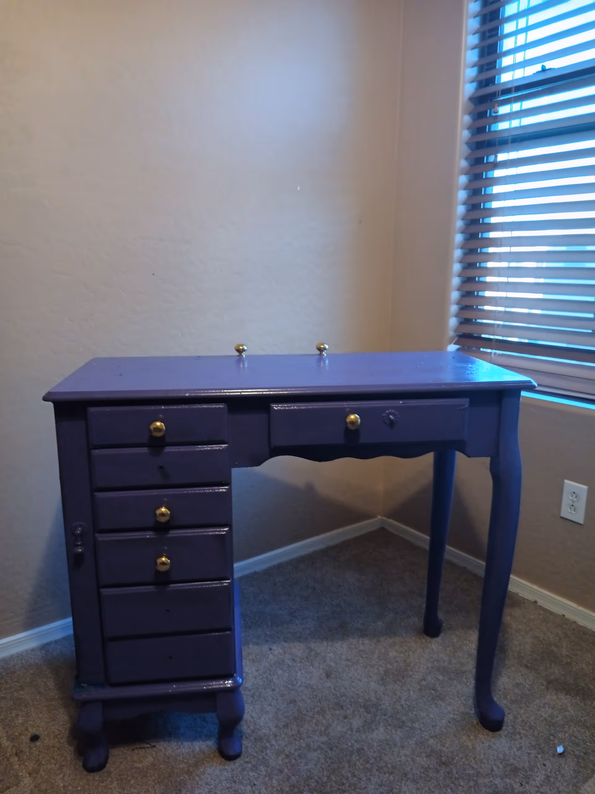 Purple wooden desk with brass knobs sits in a corner by a window.