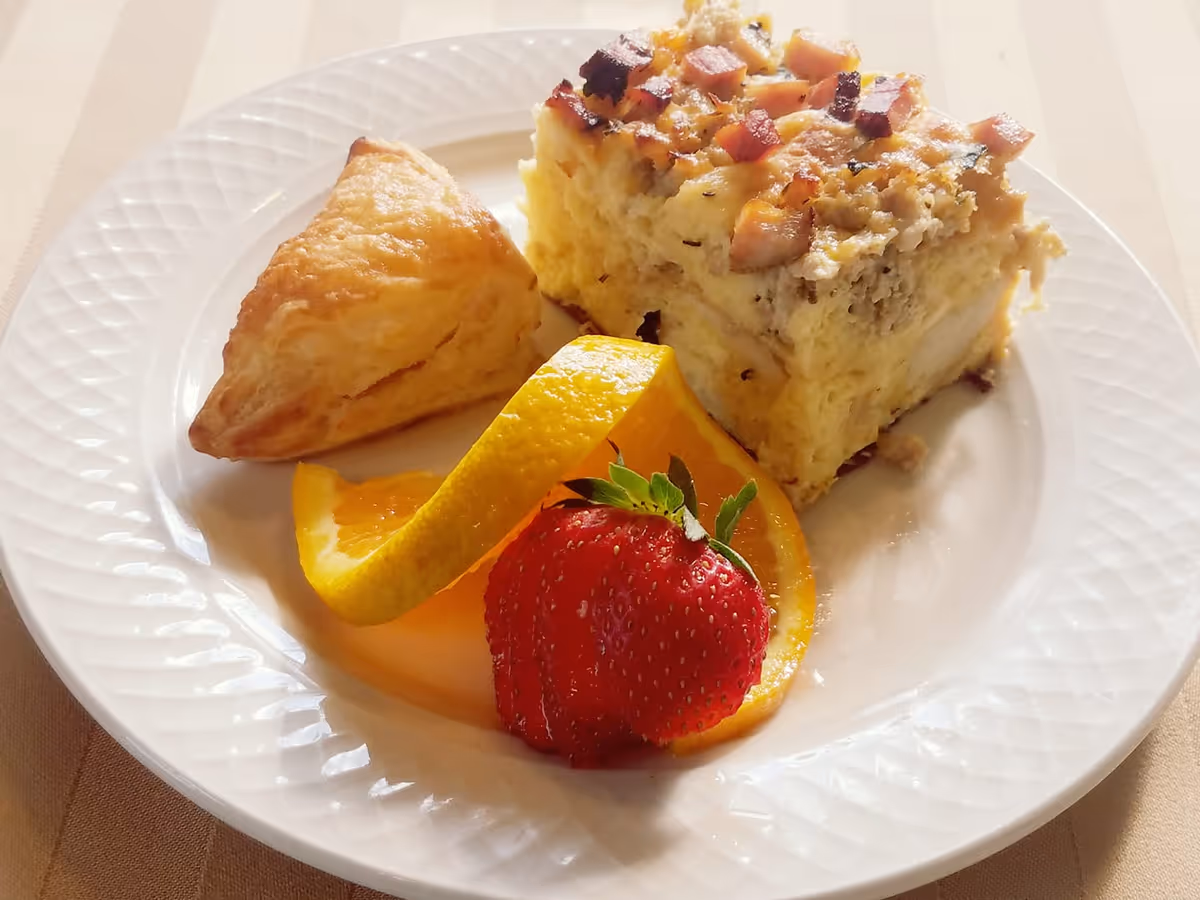 A white plate with a square egg casserole topped with diced ham, a triangular pastry, orange slices, and a strawberry.