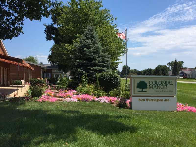 Outdoor view of a senior care facility with a well-maintained lawn, colorful flower beds, evergreen trees, and an American flag on a flagpole. A sign in the foreground reads 'COLONIAL MANOR Therapy & Senior Care 620 Warrington Ave.'