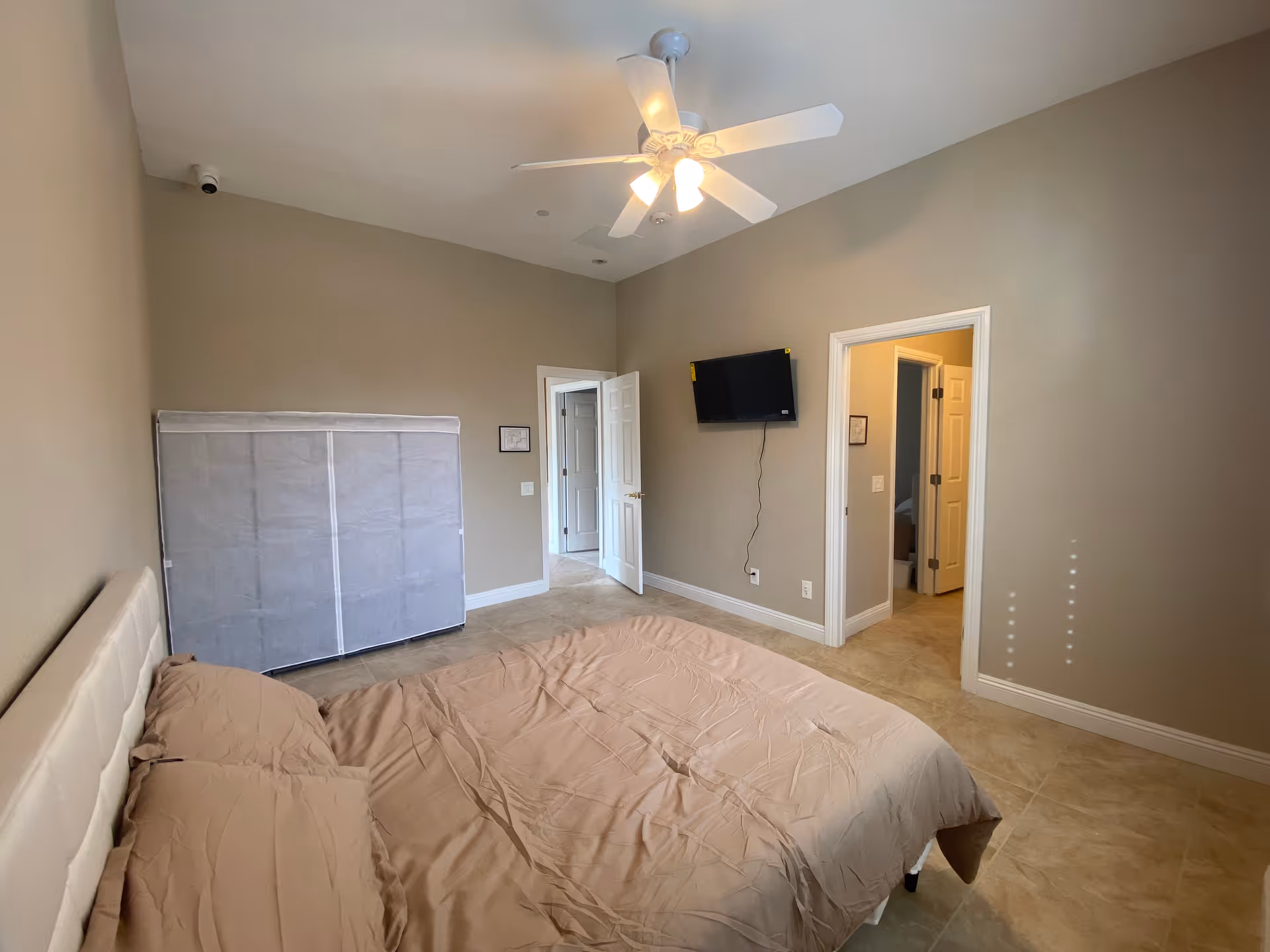 A spacious neutral-toned bedroom with a bed, ceiling fan, wardrobe, and wall-mounted TV.