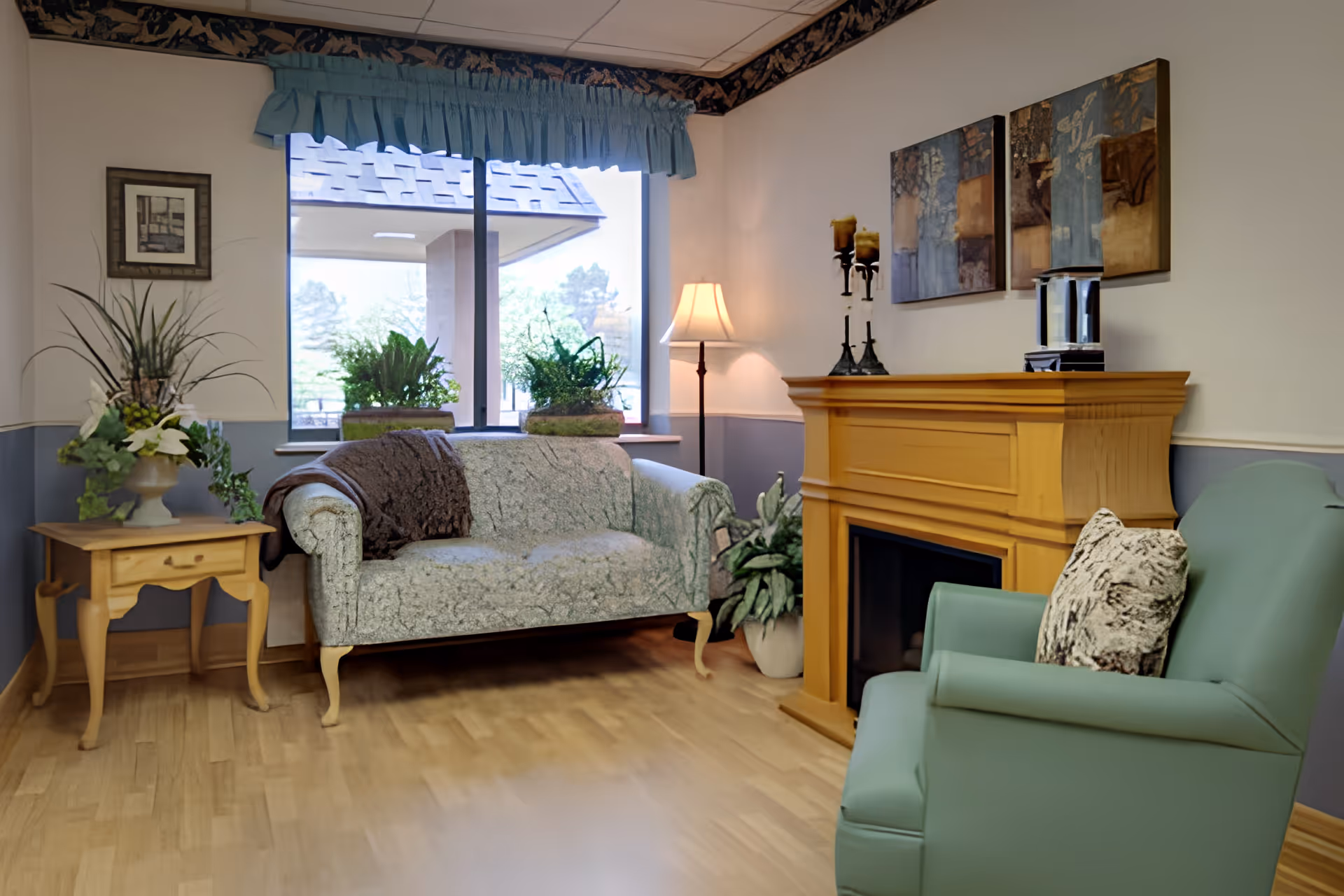 A cozy sitting area with a patterned loveseat, green armchair, wooden mantel fireplace, side table with plants, and a window with a valance.