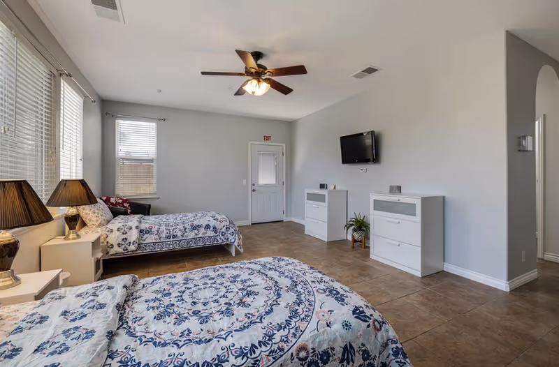 Bright shared bedroom with two twin beds, dressers, lamps, and a wall-mounted TV.