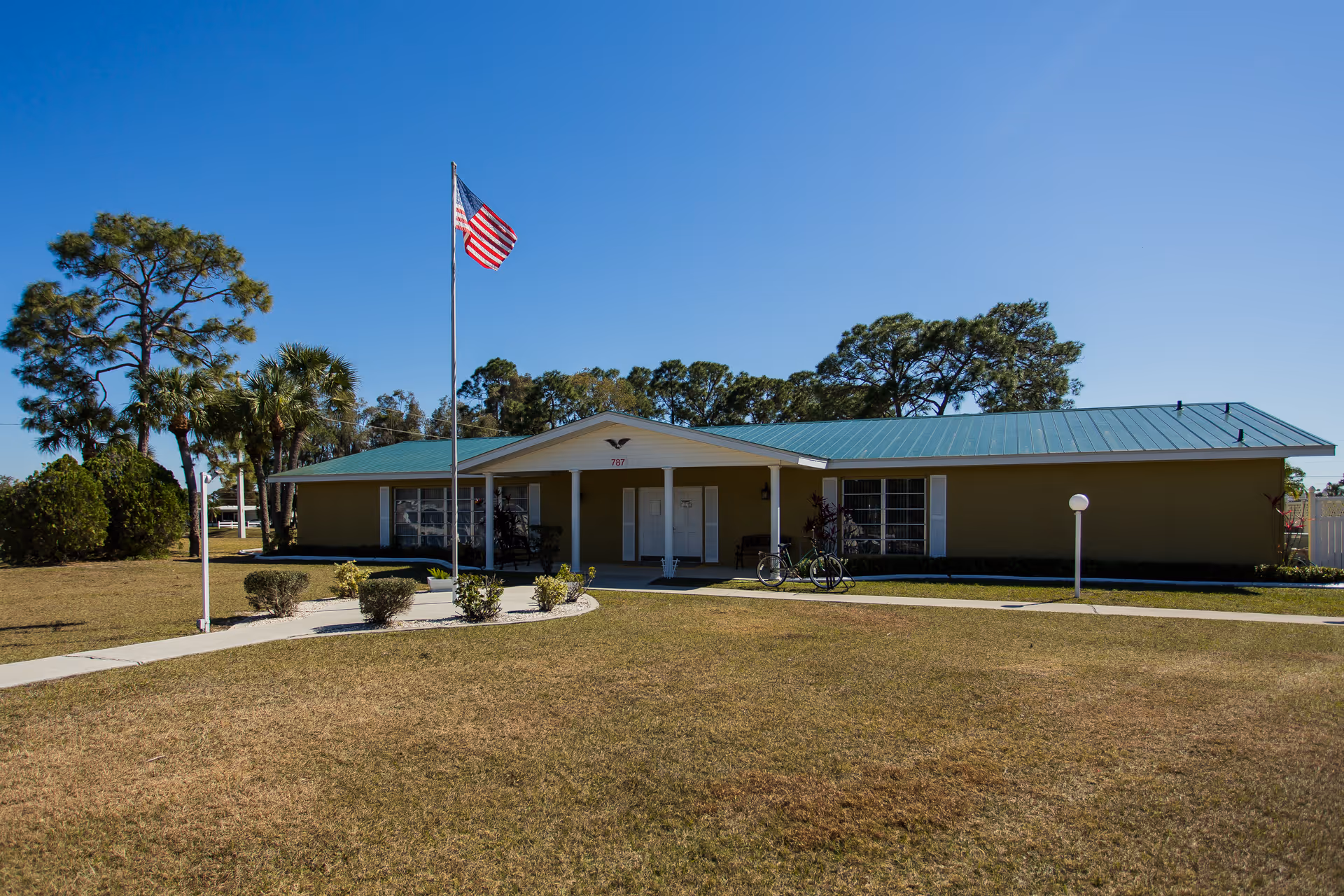 Single-story building with a green metal roof and beige walls, surrounded by a lawn and trees. An American flag flies on a flagpole in front of the building. There is a paved walkway leading to the entrance, which has a covered porch area with white double doors. Two bicycles are parked near the entrance.