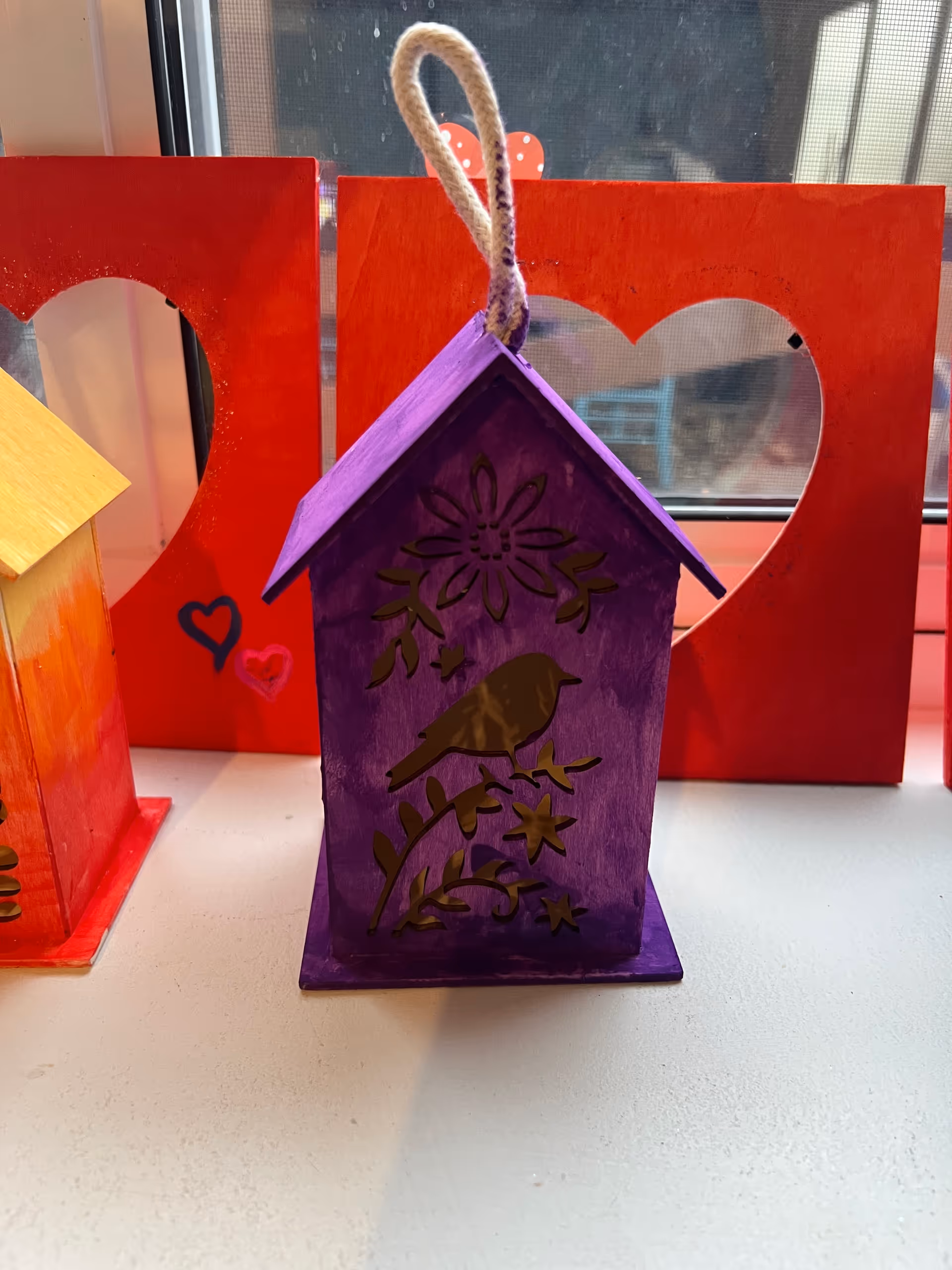 Colorful handcrafted birdhouse and heart-shaped frames on a windowsill indoors.