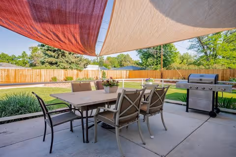 Outdoor patio area with a rectangular table surrounded by six chairs, shaded by two large fabric canopies in red and beige. A stainless steel grill is positioned nearby on the concrete patio. The background features a wooden fence, green grass, and trees under a clear blue sky.