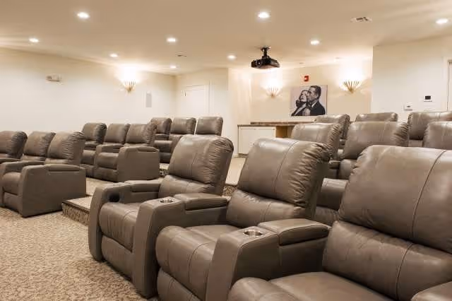 A cozy theater room with multiple rows of plush, gray reclining chairs arranged on carpeted tiered flooring. The walls are painted white and decorated with wall sconces and a black and white framed picture. A ceiling-mounted projector is visible at the center of the ceiling.