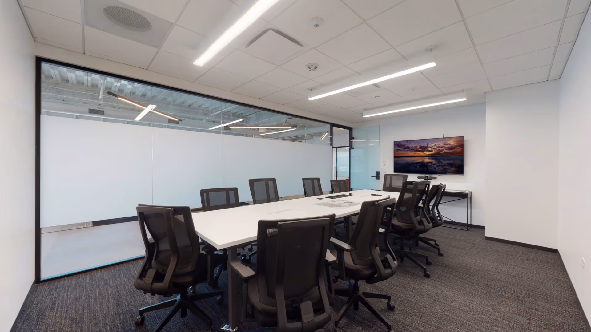 Glass-walled conference room with a long white table, black chairs, and a wall-mounted screen.