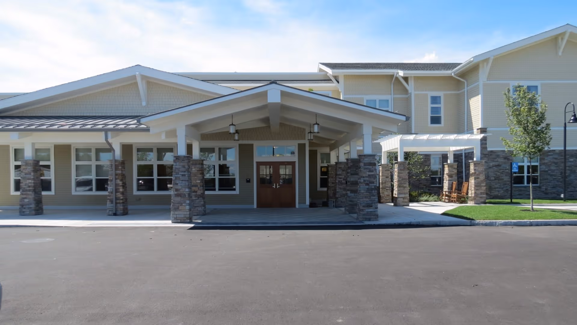 Front exterior view of a senior living facility building with a covered entrance supported by stone pillars, multiple windows, a small tree, and a paved driveway in front.