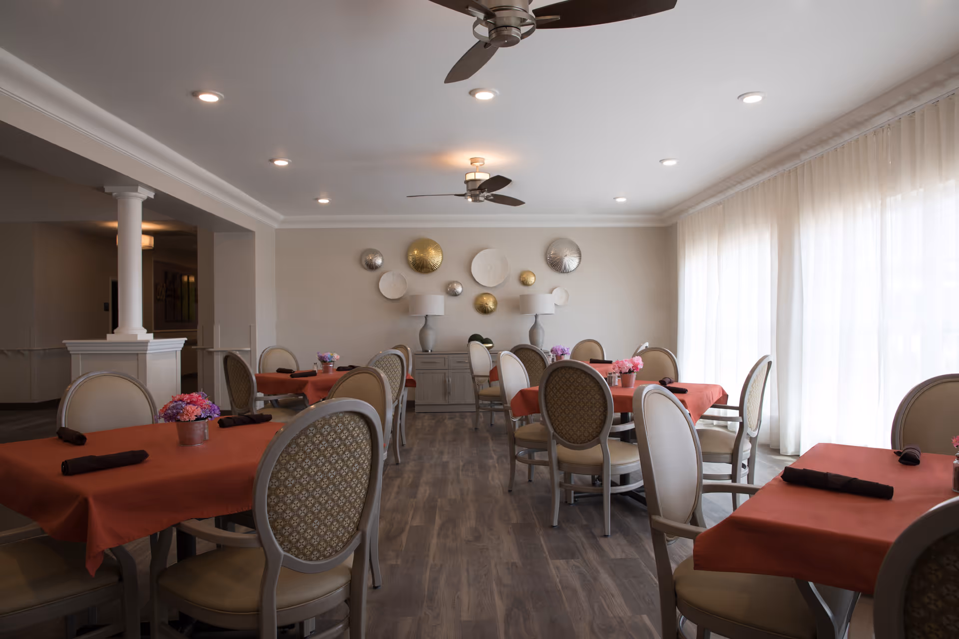 A dining room with several tables covered in red tablecloths, each set with black napkins and small flower arrangements. The room has upholstered chairs with patterned backs, wood flooring, ceiling fans, recessed lighting, and large windows with sheer white curtains allowing natural light to fill the space. Decorative circular wall art and two table lamps are visible on a sideboard against the far wall.