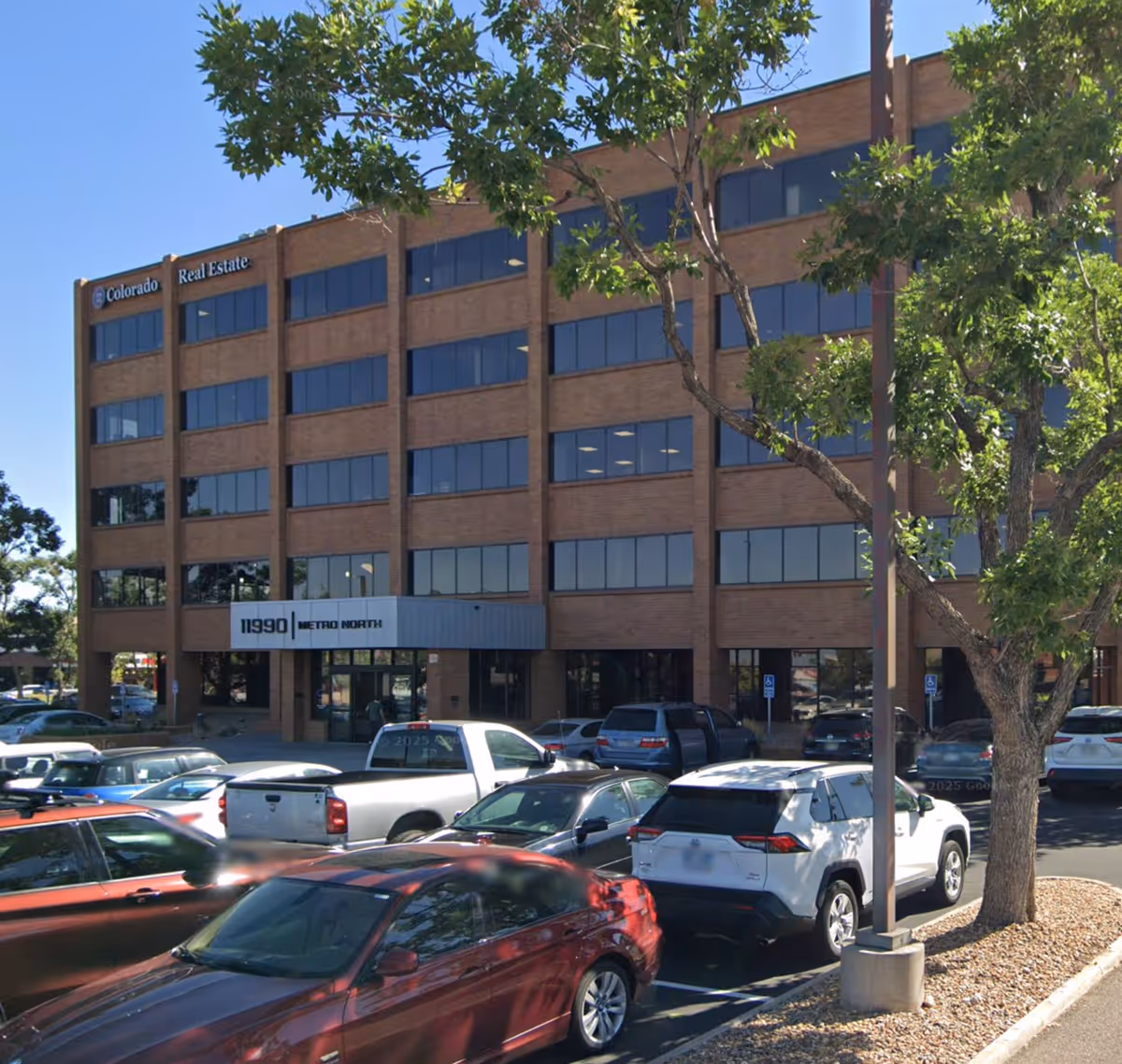 Brick office building with cars in the parking lot and trees in front.