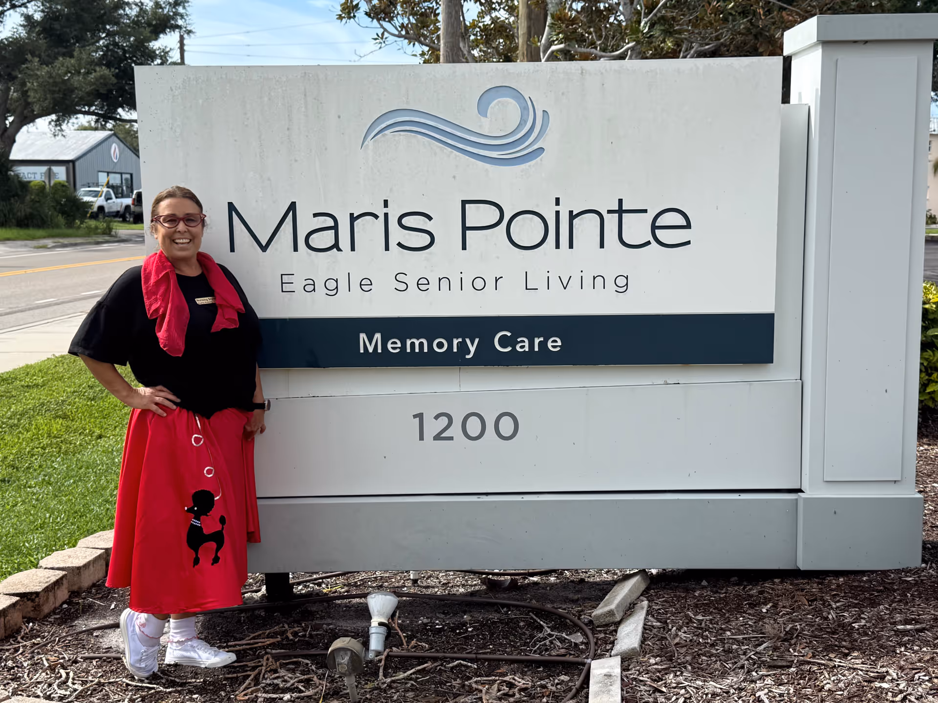 A woman in a red skirt stands beside a sign for Maris Pointe Eagle Senior Living.