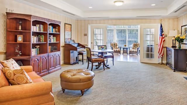 Spacious senior living common area with sofa, bookshelf, piano, seating, glass doors, and American flag.