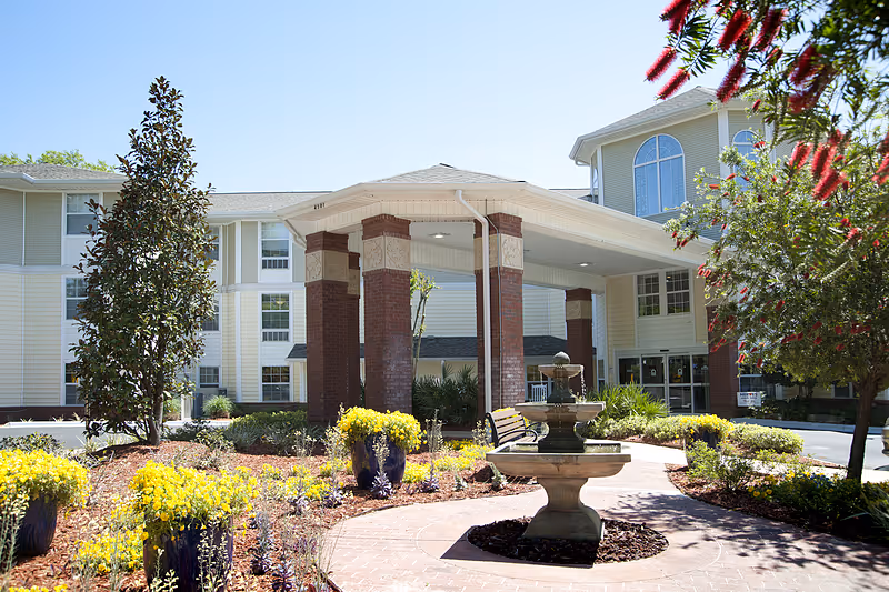 Exterior view of Allegro Senior Living in Tallahassee, FL, showing the entrance with a covered driveway supported by brick columns, surrounded by landscaped gardens with yellow flowers, a water fountain, and trees under a clear blue sky.