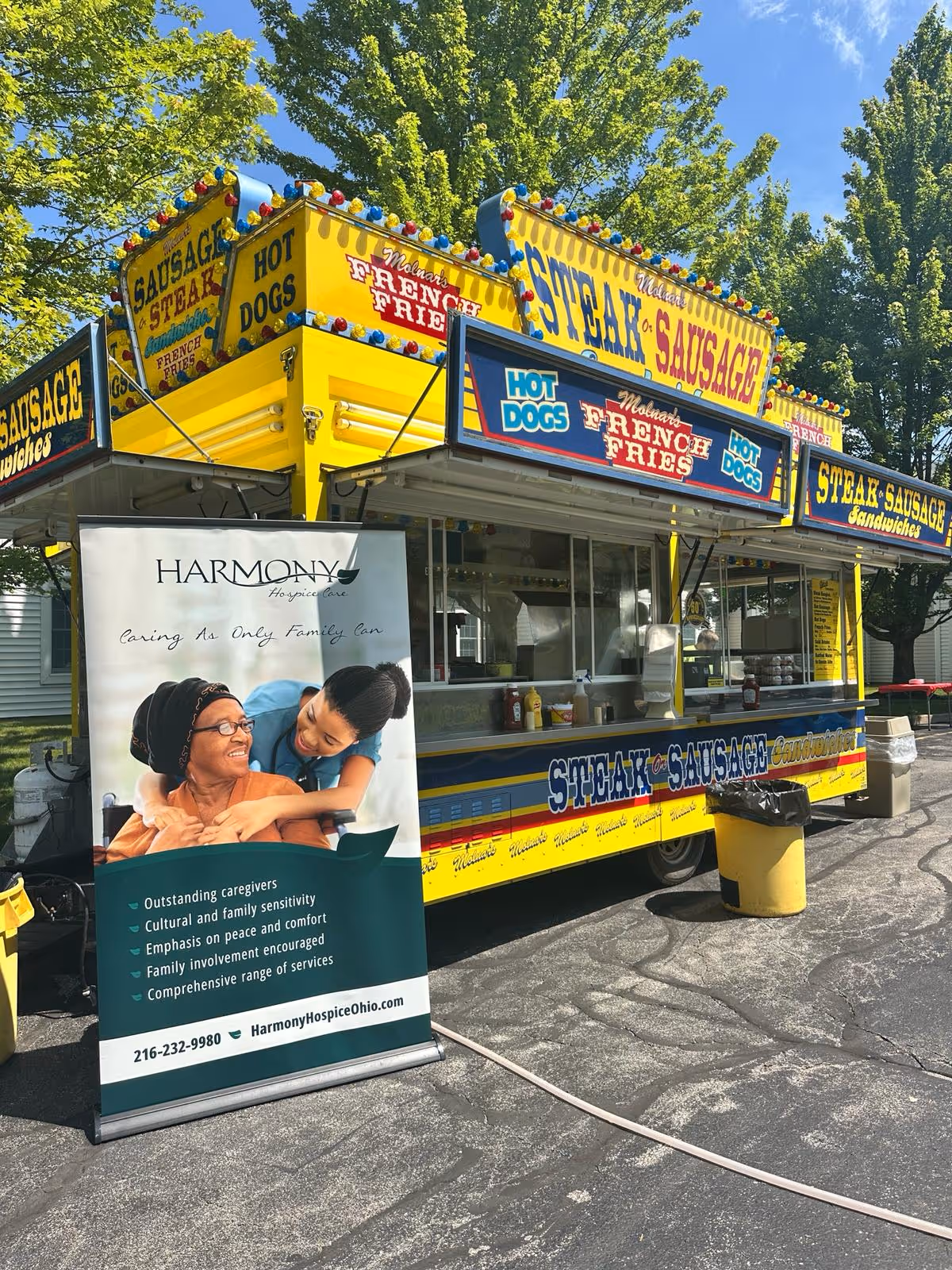 Bright yellow food truck with sausage signs beside Harmony Hospice banner