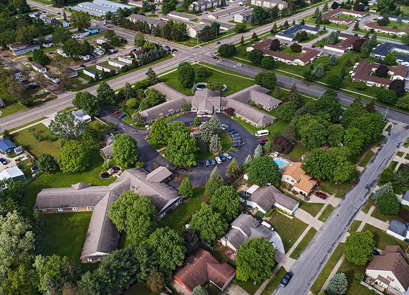Aerial view of the American House Kentwood senior living facility surrounded by trees, roads, and residential houses. The facility buildings are arranged in a U-shape with a parking lot in the center, and there is a swimming pool visible near one of the buildings.