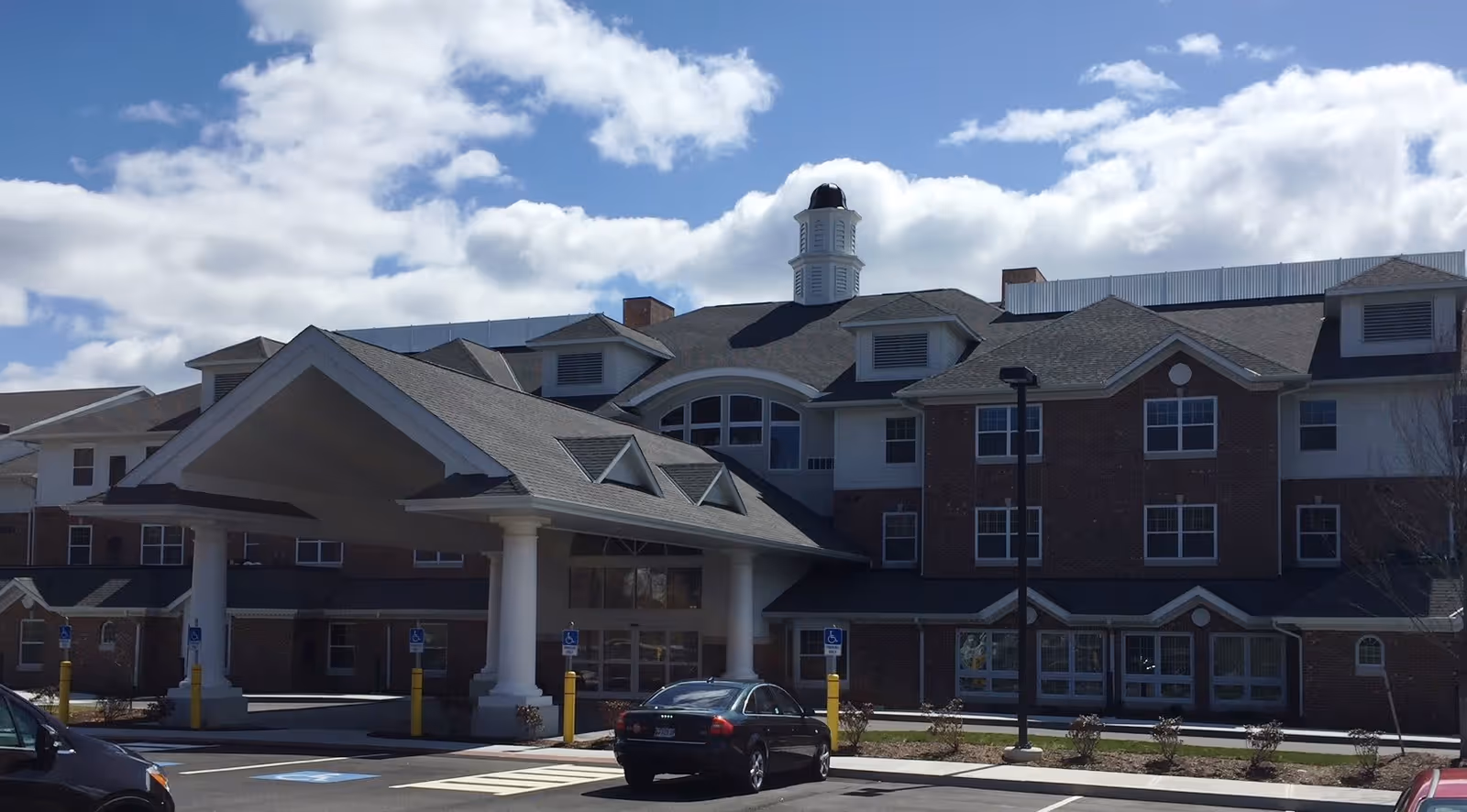 Exterior view of a multi-story brick building with a large covered entrance supported by white columns, several windows, and a cupola on the roof under a partly cloudy sky. Cars are parked in front of the building in a parking lot.