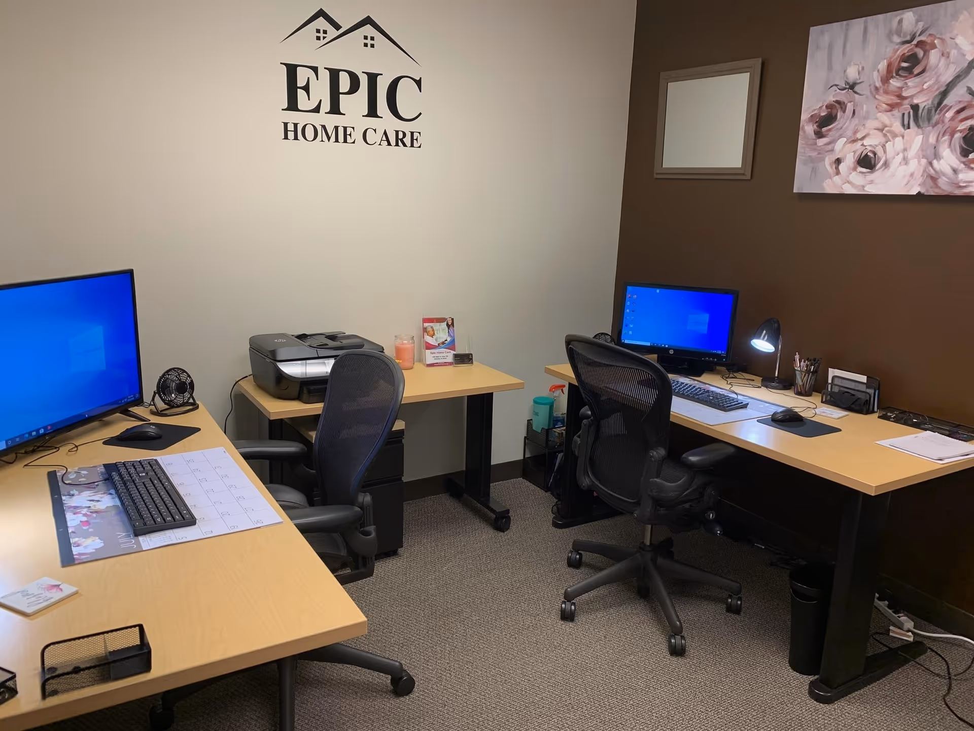Office workspace with desks, computers, printer, and Epic Home Care logo on wall.