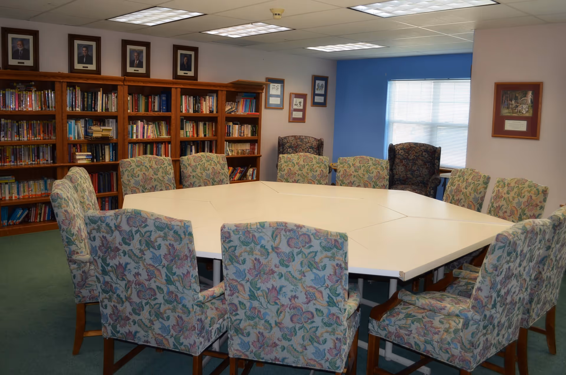 A bright interior meeting room with a large white hexagonal table surrounded by floral upholstered chairs and bookshelves.