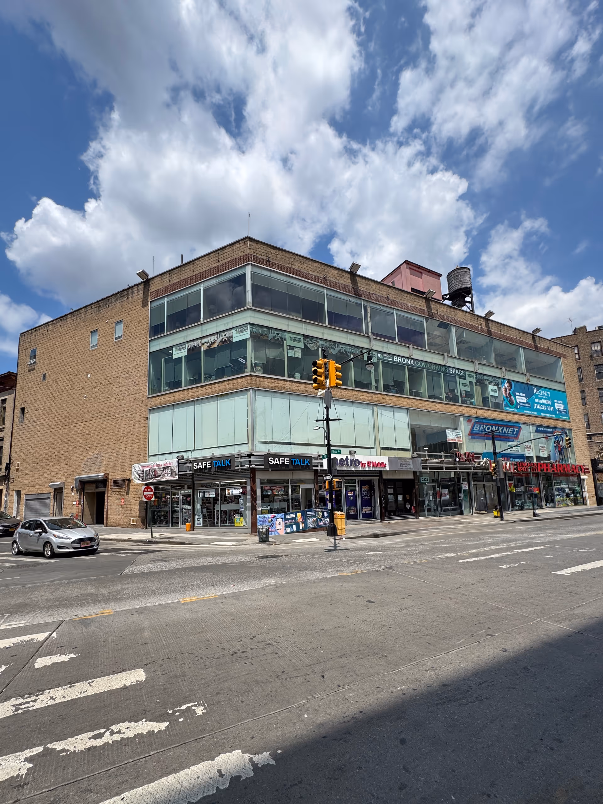 Corner street view of a brick, glass-front building with storefronts and a crosswalk.