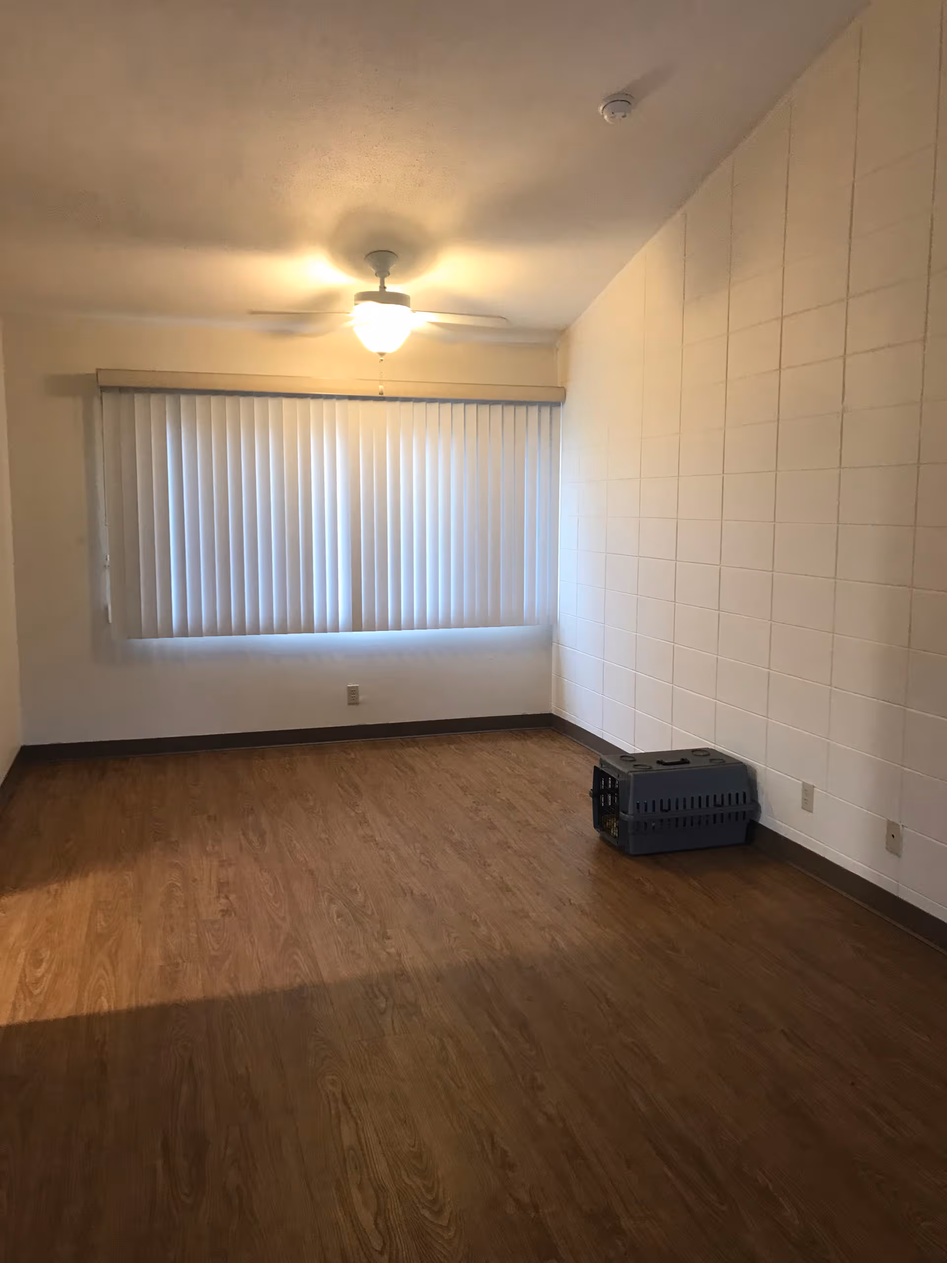 Empty interior room with blinds, ceiling fan, wood floor, and a pet crate.