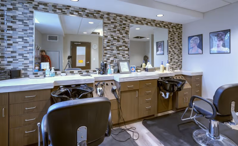 Hair salon station with two styling chairs, black shampoo basins, and a large mirror over a mosaic-tiled backsplash.