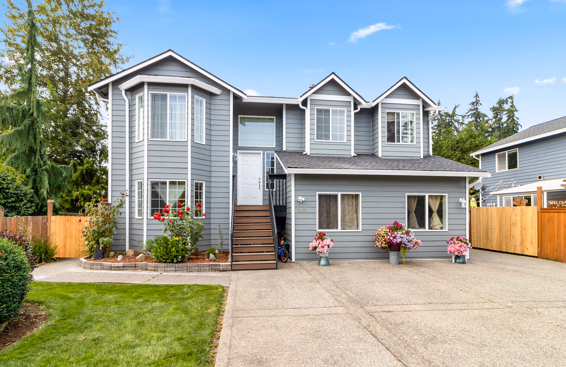 Gray two-story home with front stairs, lawn, and potted flowers, driveway, and wooden fencing.