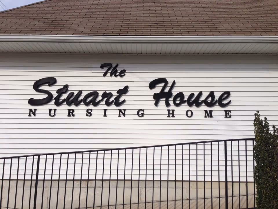 Exterior wall of a building with a sign that reads 'The Stuart House Nursing Home' in black letters on white siding, with a brown shingled roof above and a black metal railing in front.
