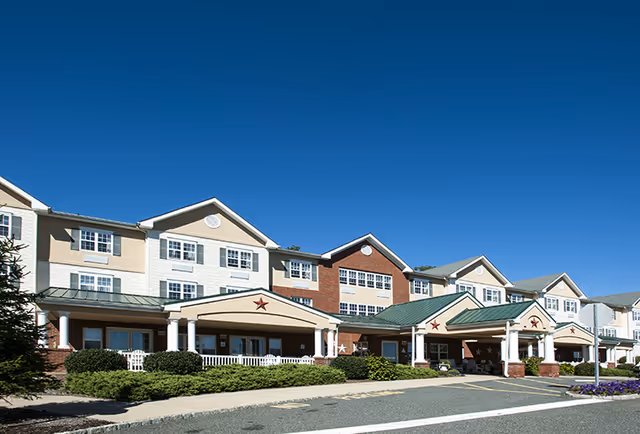 Front entrance of a multi-story senior living building with covered porticos, white columns and green roofs under a clear blue sky.