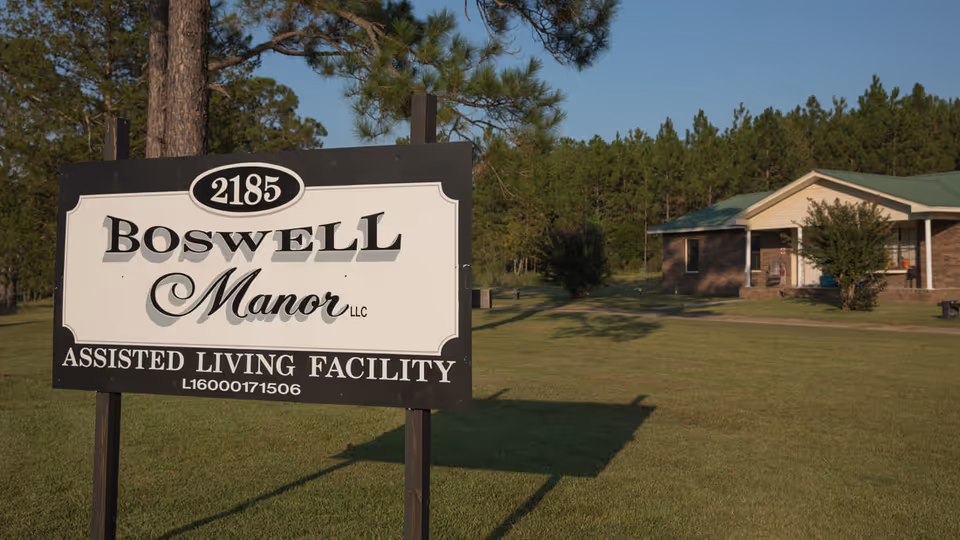 A large yard with a sign reading "Boswell Manor LLC Assisted Living Facility" in front of a one-story building and trees.