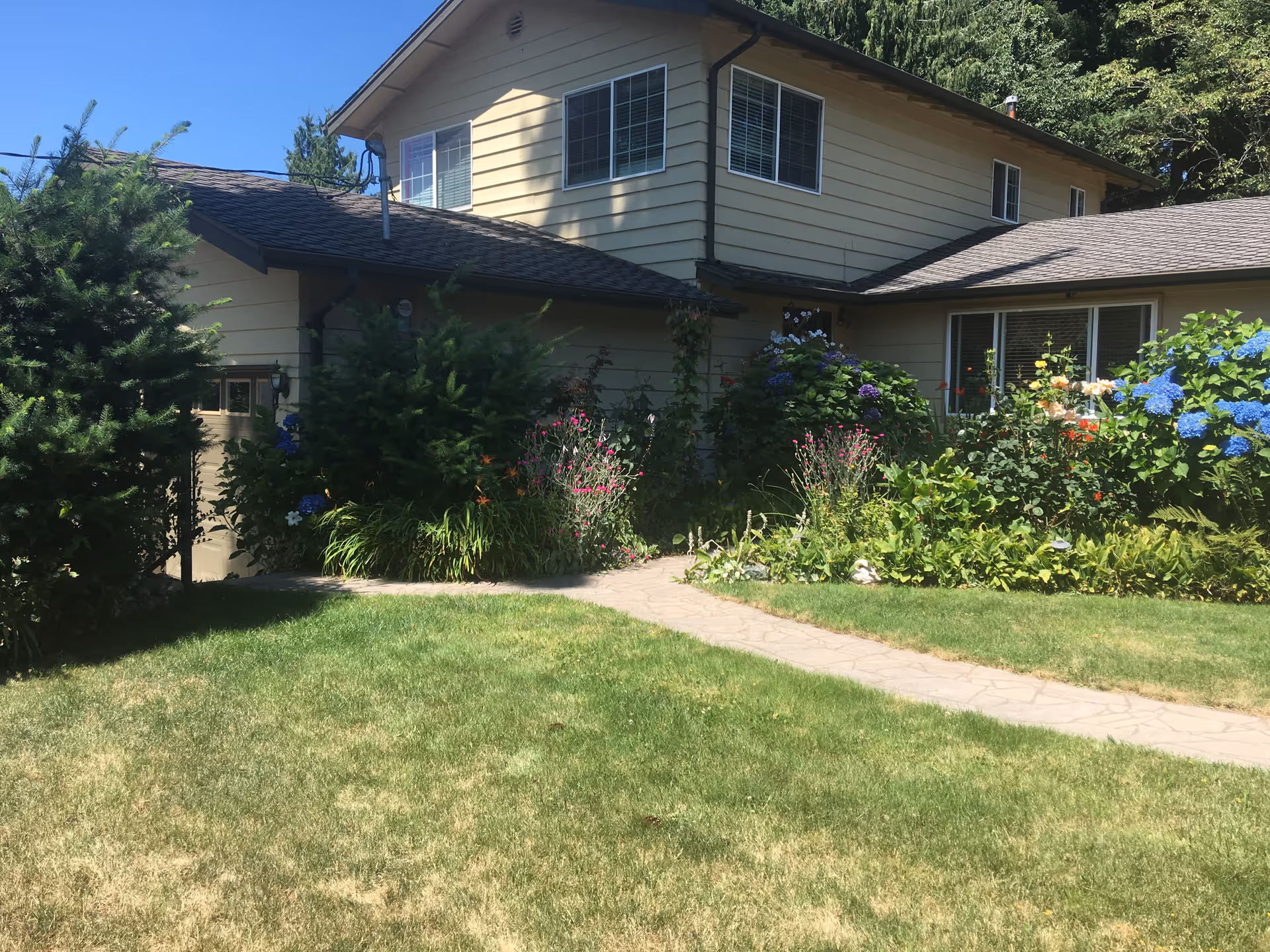Two-story beige house with a lush garden, stone walkway, and vibrant flowers.