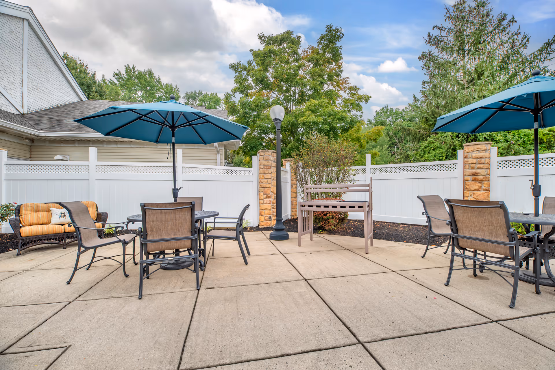 Outdoor patio with multiple tables, chairs and blue umbrellas on a paved courtyard surrounded by a white fence and trees.