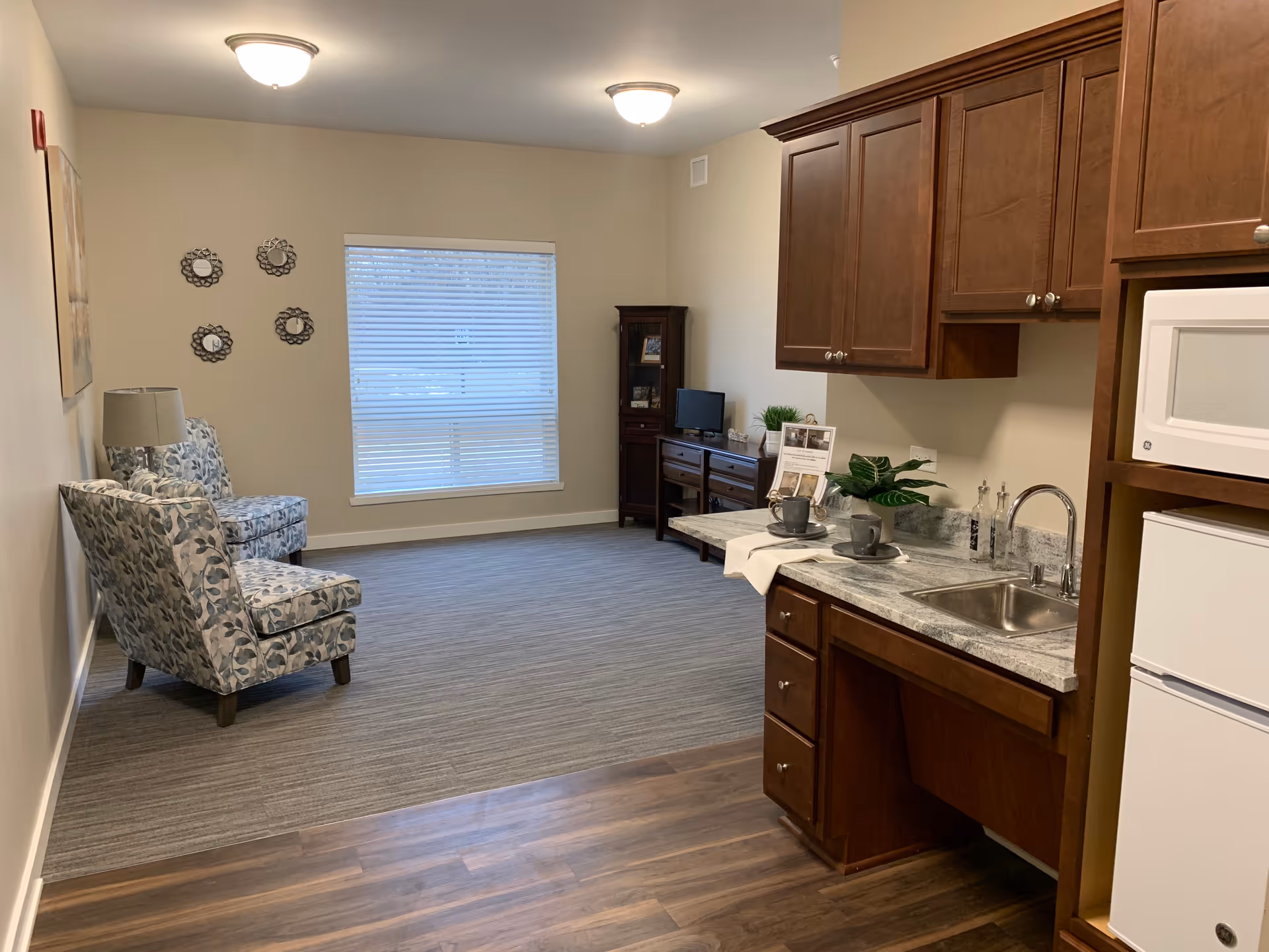 A cozy living area with two patterned armchairs, a lamp, and decorative wall mirrors on the left side. A window with blinds is centered on the back wall. On the right side, there is a kitchenette with wooden cabinets, a small sink, a microwave, and a mini refrigerator. The floor transitions from wood near the kitchenette to carpet in the seating area.