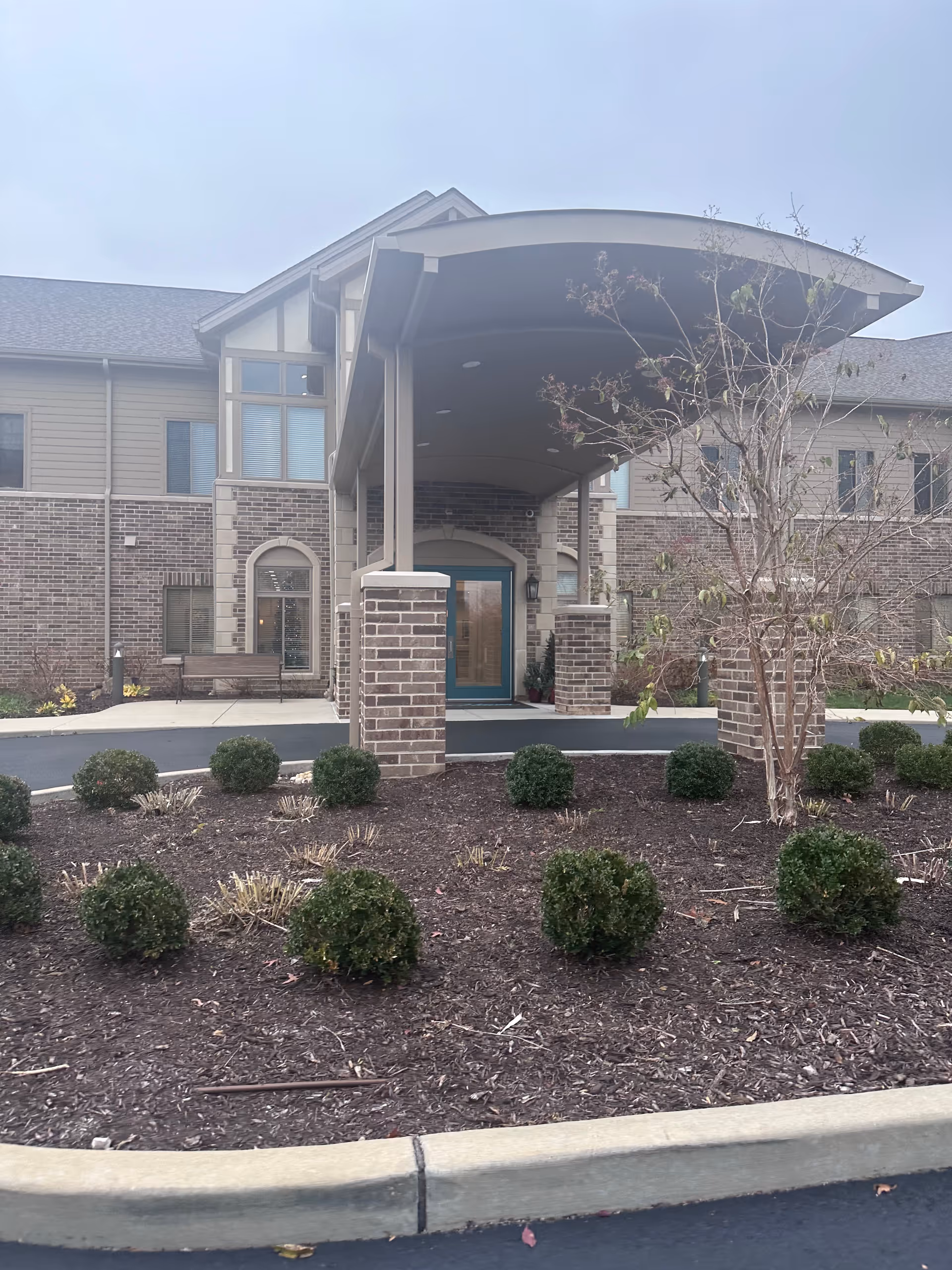 Front entrance of a two-story brick building with a covered porte-cochere and landscaped shrub beds.