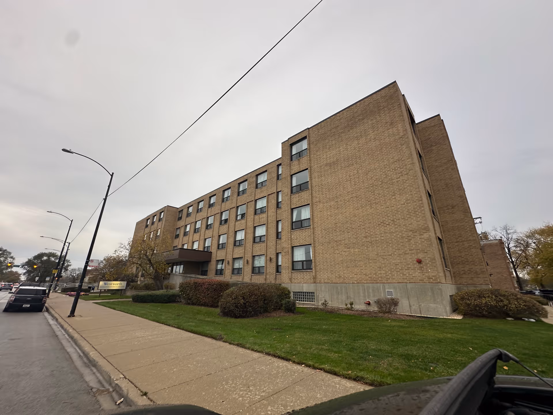 Multi-story brick healthcare building seen from the street with a sidewalk, lawn, and parked cars in the foreground.