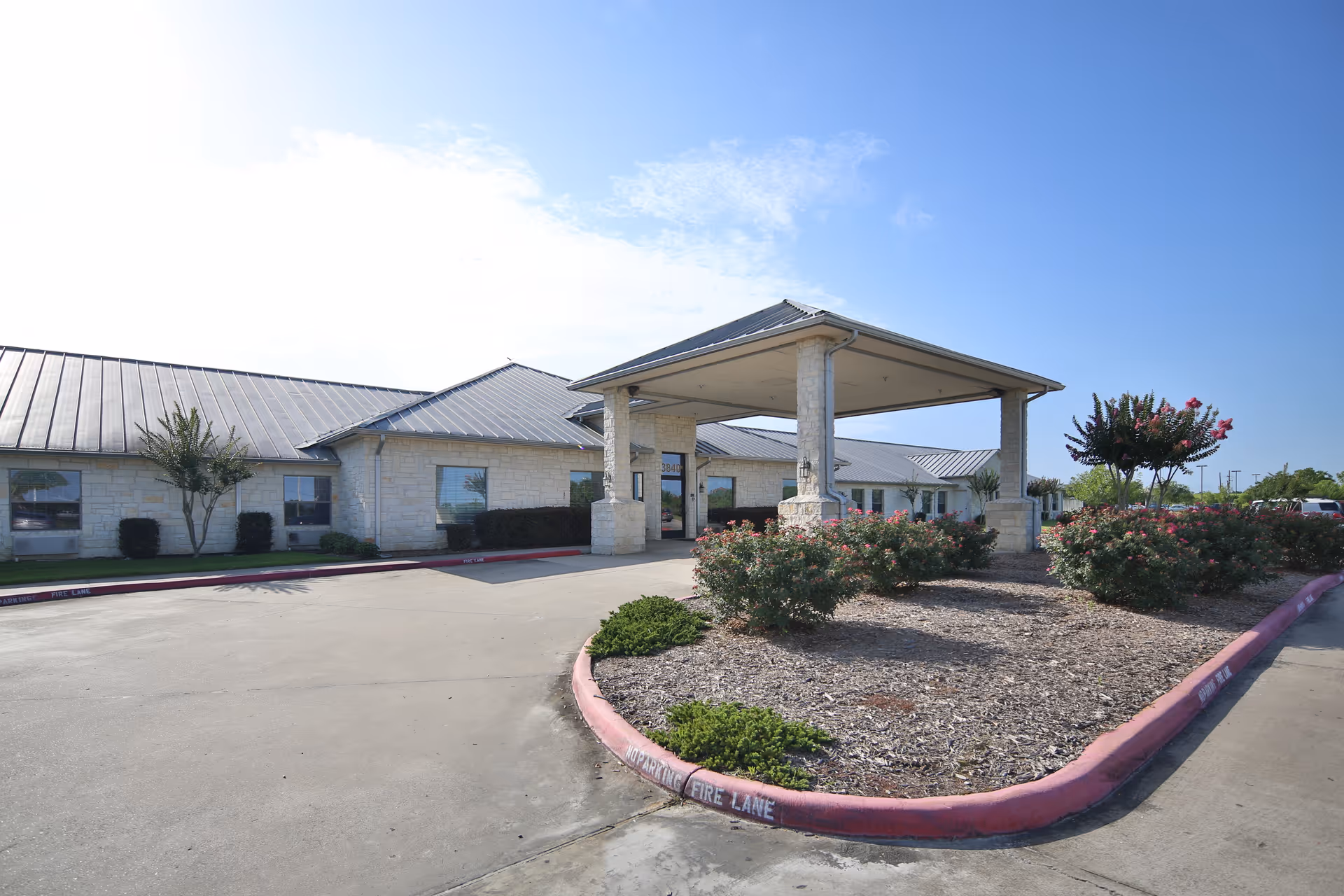 Exterior view of Jefferson Nursing and Rehabilitation Center showing a single-story building with a metal roof, a covered entrance supported by stone pillars, landscaped bushes and trees, and a driveway with a fire lane marked in red.