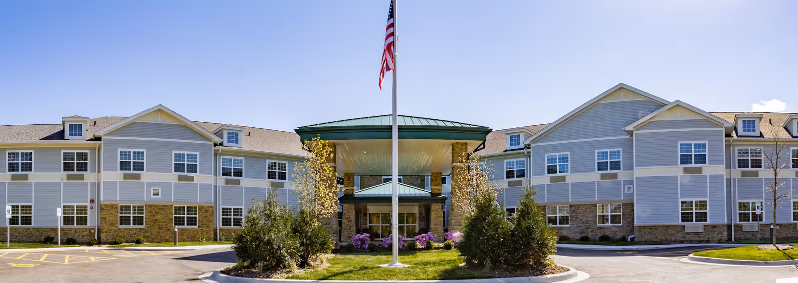 Front exterior view of a senior living facility with a central entrance covered by a green roof. The building has light gray siding with stone accents on the lower part and multiple windows. There is a flagpole with an American flag in front of the entrance, surrounded by landscaping with bushes and flowers under a clear blue sky.