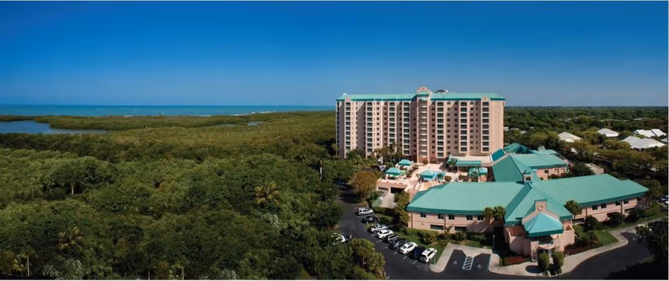 Aerial view of Premier Place facility surrounded by dense greenery with a large multi-story building and several smaller buildings with green roofs. There is a parking lot with cars and a clear blue sky above.