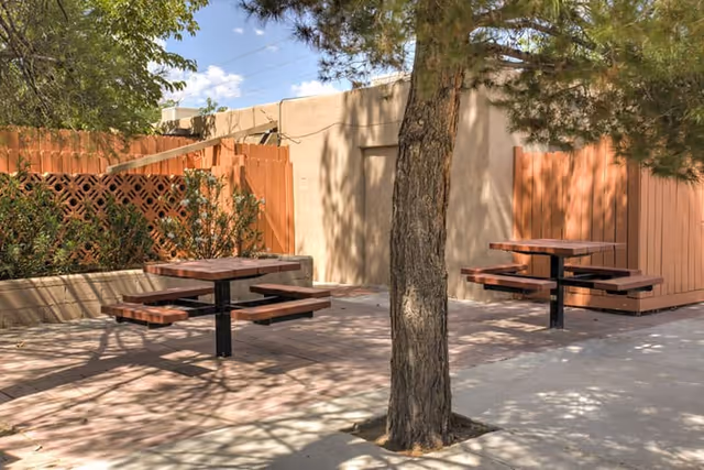 Outdoor seating area with two wooden picnic tables with attached benches on a concrete surface, surrounded by a wooden fence and some greenery, with a tree in the foreground casting shadows.
