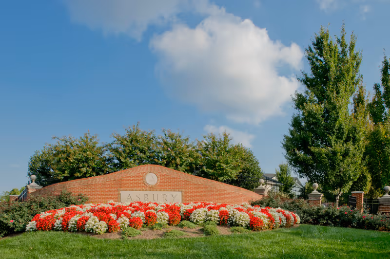 Brick entrance sign reading 'Asbury' surrounded by red and white flowers, green lawn and trees under a blue sky.