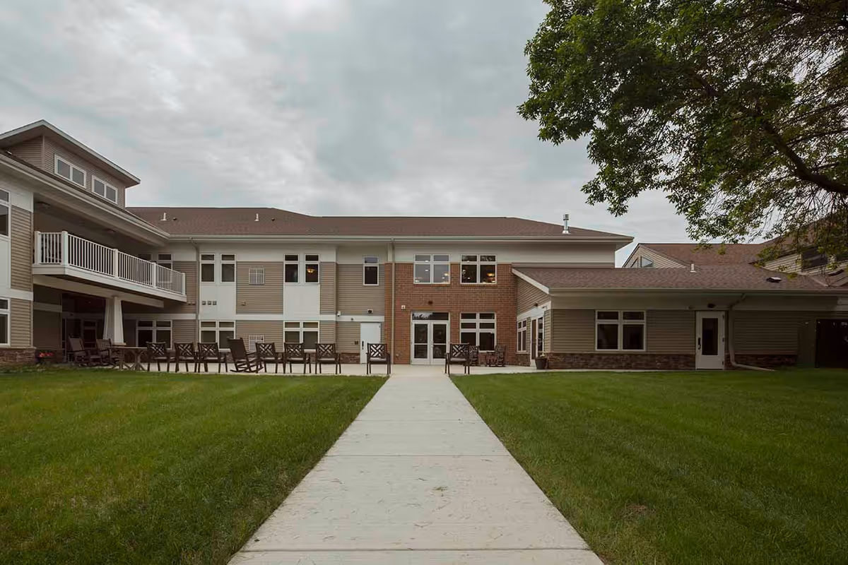Exterior view of Melrose Park View Place, showing a two-story building with beige and brick facade, multiple windows, and a concrete walkway leading to the entrance. There are several chairs arranged on the patio area, and a large tree is visible on the right side of the image under a cloudy sky.