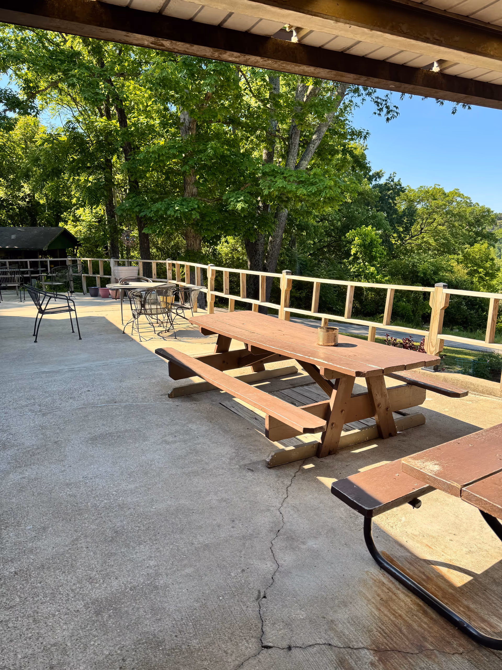 Sunny outdoor terrace with wooden picnic tables, metal chairs, and surrounding green trees.