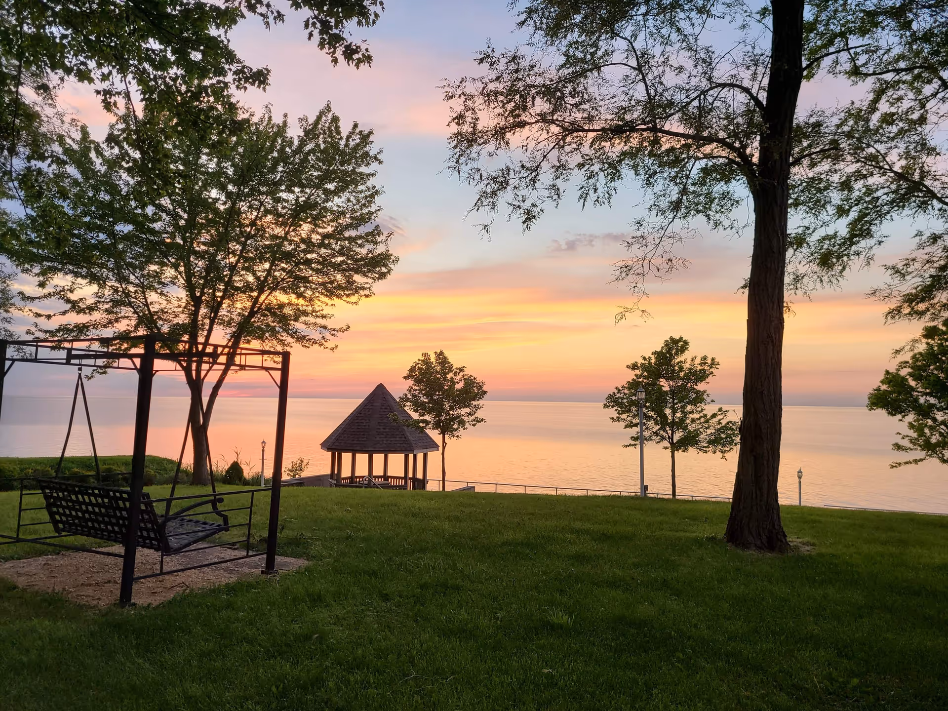 Sunset over a lakeside lawn with a gazebo, trees, and a bench swing near the lake.